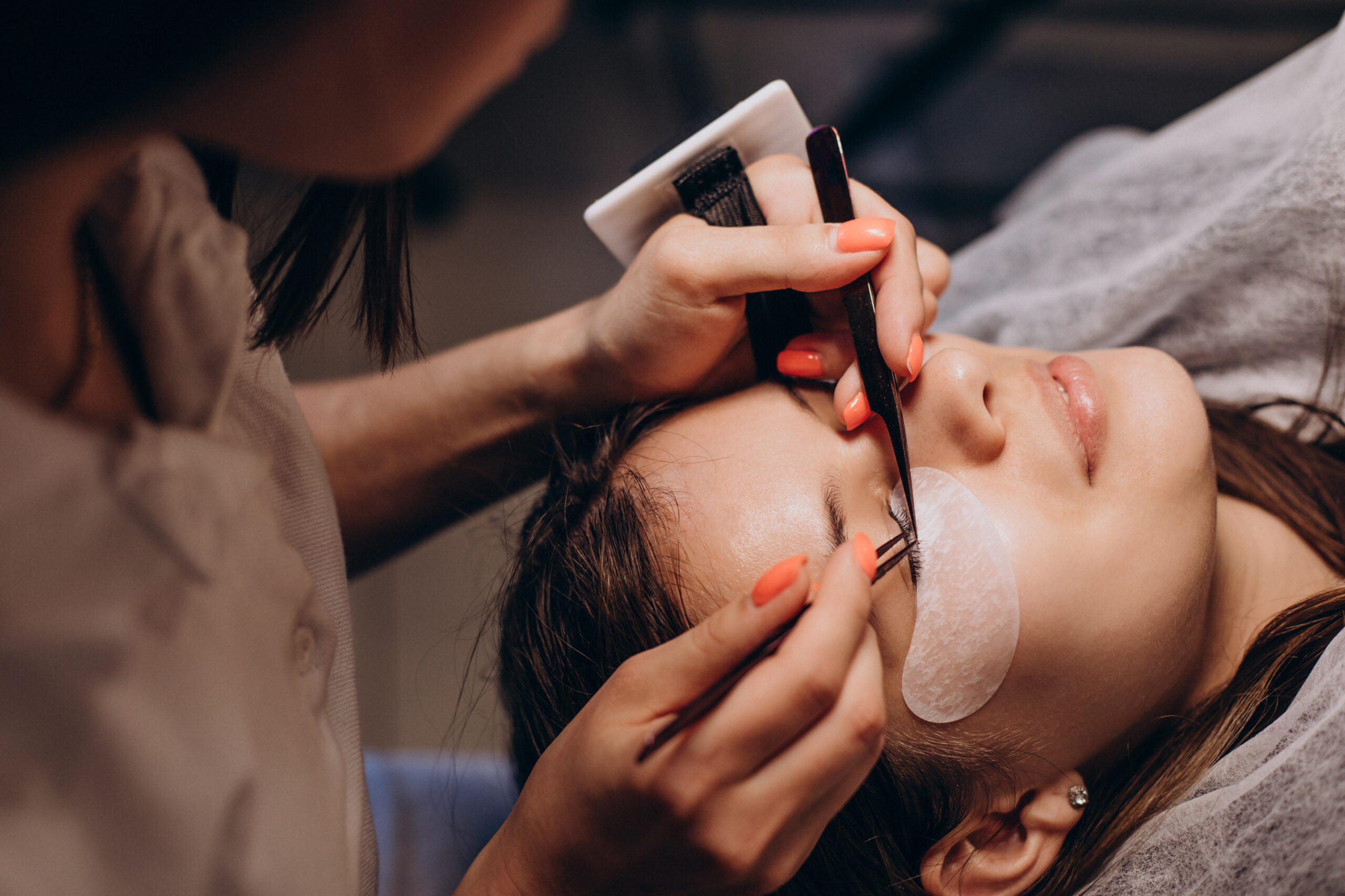 A woman receiving eyebrow shaping from a professional, showcasing various eyelash extension styles in the background.